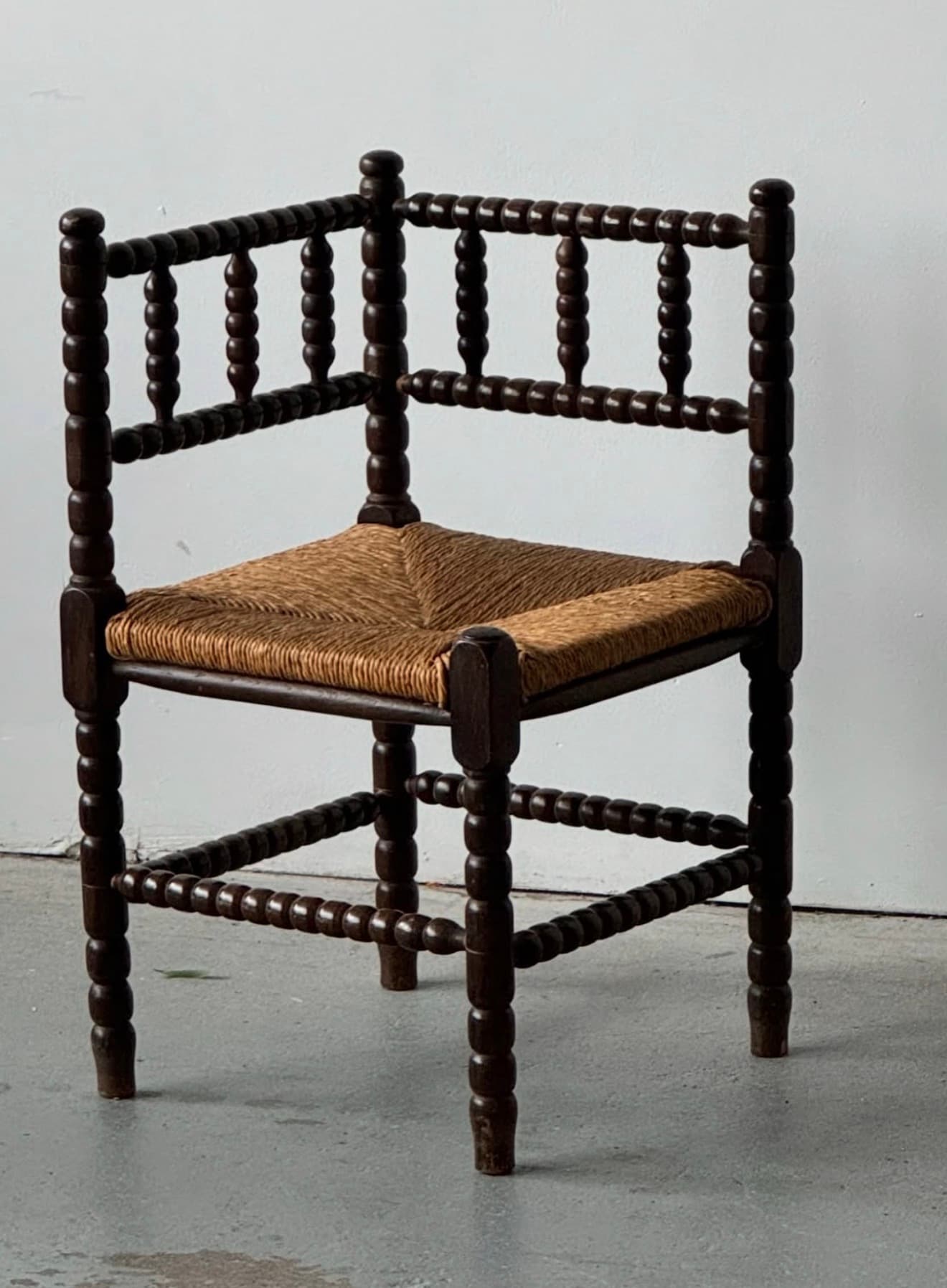A dark brown stained wooden corner chair featuring extensive bobbin turning on its frame, backrest, and legs. The seat is constructed from tightly woven natural rush material, providing a textured contrast to the smooth, turned wood. Its unique corner-fitting design suggests placement in a room's angle. — 3D model