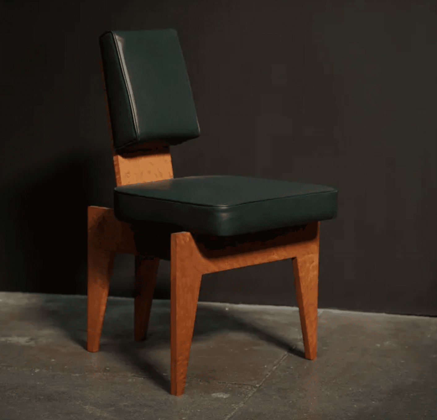 An armless chair featuring a distinctive geometric wooden frame with a light-colored, possibly burled, wood grain. The seat and separate backrest are upholstered in a dark green material, likely leather or a high-quality synthetic, offering a plush appearance. The wooden base is composed of thick, angular planks forming the legs and supporting structure, with a splayed design that tapers slightly towards the floor. The chair has a low profile backrest, suggesting an accent or side chair function. — 3D model