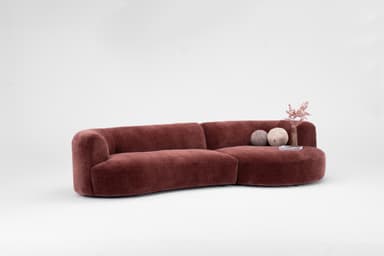 A large, contemporary curved sofa upholstered in a rich, burnt orange velvet or similar plush fabric. The sofa features a continuous, undulating form with a low backrest and seamlessly integrated armrests. Its design is characterized by soft, rounded edges and a low-profile base, giving it a sleek and inviting appearance. Decorative pillows and a vase with dried botanicals are placed on one end of the sofa. — 3D model