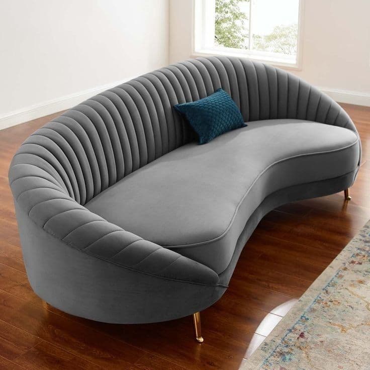 A contemporary curved loveseat featuring a light grey velvet upholstery with a distinctive channel-tufted backrest and armrests. It has a single, elongated seat cushion and is supported by slender, tapered golden metal legs. A small teal accent pillow is included. — 3D model
