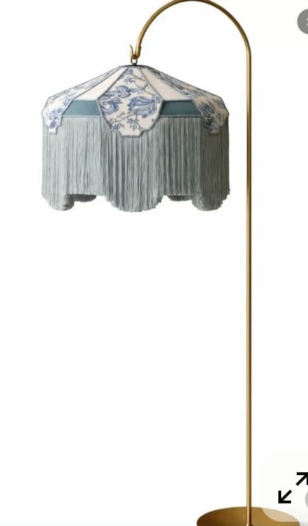 A floor lamp featuring a decorative lampshade with a blue floral pattern on a white background, accented with a teal band and a long, pale blue fringe. The lampshade is supported by a curved, slender brass arm extending from a thin brass pole, which stands on a round brass base. — 3D model