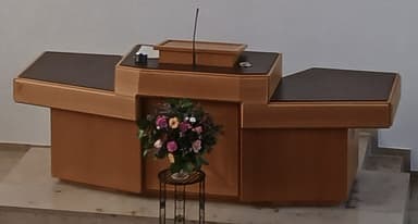 A tiered wooden reception desk with dark brown faux leather inlays on its surfaces, featuring an integrated podium with a gooseneck microphone. A small black metal plant stand with a vase of flowers is placed next to it. — 3D model