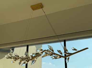 A decorative chandelier designed to resemble a branch with leaves. It features a matte gold-toned metal frame with a branching structure. Attached to the branches are multiple clear or frosted glass diffusers shaped like elongated, faceted crystals or buds, each housing a small light bulb. The chandelier is suspended by thin wires from a rectangular ceiling canopy. — 3D model