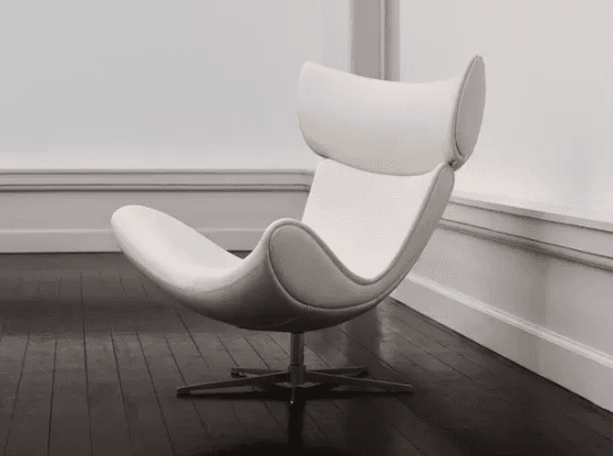 A modern, ergonomic lounge chair with a distinctive high-back design. The chair features a curved seat and backrest, upholstered in a smooth, white material. It is supported by a central pedestal base with four splayed legs, allowing for swivel functionality. The chair is positioned on a dark wooden floor against a white wall with molding. — 3D model