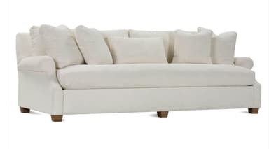 A contemporary sofa featuring a clean, off-white fabric upholstery, rolled arms, and multiple loose back cushions. The sofa sits on short, exposed wooden block feet. It includes several accent pillows, suggesting a comfortable and relaxed aesthetic. — 3D model