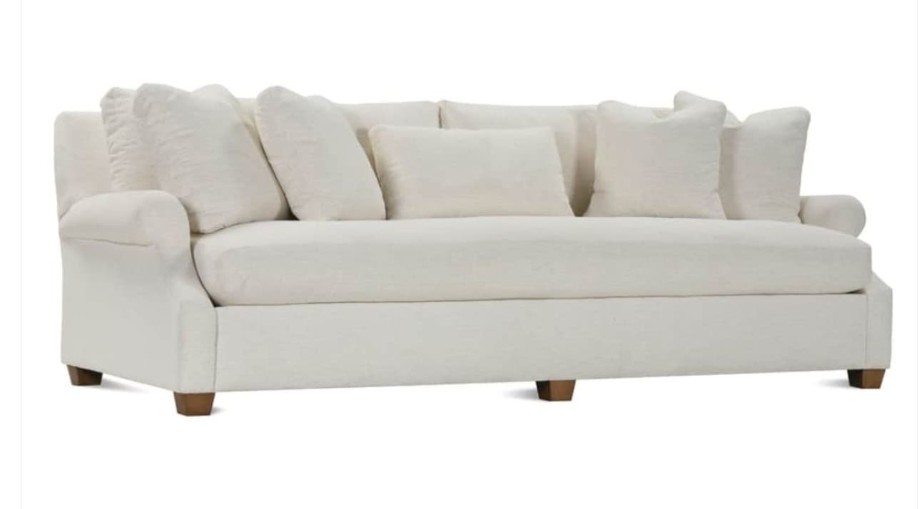 A contemporary sofa featuring a clean, off-white fabric upholstery, rolled arms, and multiple loose back cushions. The sofa sits on short, exposed wooden block feet. It includes several accent pillows, suggesting a comfortable and relaxed aesthetic. — 3D model