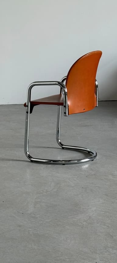 A cantilever armchair with a chrome-plated tubular steel frame and a brown leather seat and backrest. The frame has a continuous curve forming the legs, seat support, and armrests. The backrest is slightly curved and extends upwards, with a visible seam running along its edge. The seat is a flat plane that connects to the frame. The overall design suggests a mid-century modern or Bauhaus influence. — 3D model