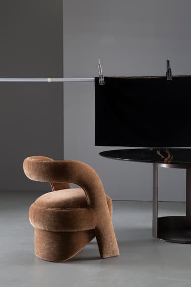 A contemporary sculptural accent chair fully upholstered in a textured medium brown fabric, featuring a continuous, curvilinear backrest and armrests that flow seamlessly into an integrated, upholstered cylindrical base. — 3D model