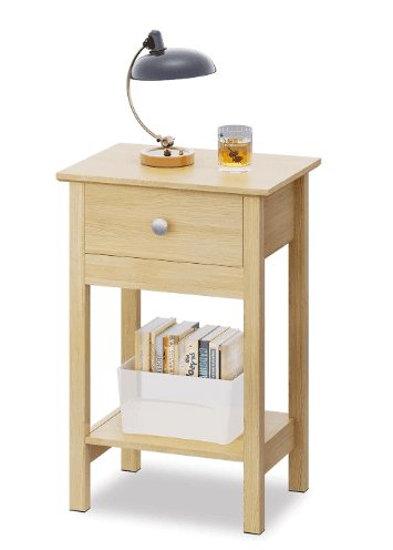 A light-colored wooden nightstand featuring a single drawer with a round silver-tone knob and an open lower shelf. The table stands on four straight, square-profile legs. A desk lamp, a glass of amber liquid, and a clear container holding books are displayed on and within the nightstand. — 3D model