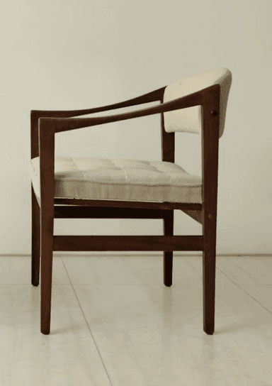 A mid-century modern armchair featuring a dark wood frame and light beige fabric upholstery. The chair has a clean, rectilinear wooden frame with subtly curved armrests. Both the seat and back are padded, with the seat cushion exhibiting button tufting, suggesting a comfortable yet structured design. The wooden legs are straight, and the overall aesthetic is minimalist and functional. — 3D model