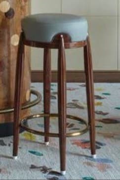 A modern bar stool with a round, upholstered seat and four tapered wooden legs. A circular metal footrest connects the legs partway down. The seat appears to be made of light gray leather or a similar material, and the frame and legs are constructed from dark wood. The footrest is a brass-toned metal ring, and the feet of the legs have white caps. — 3D model