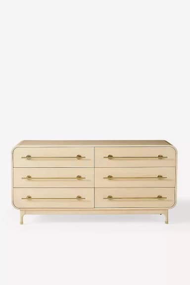 A modern six-drawer dresser featuring a light wood finish and elegant brass bar pulls. The piece has soft, rounded edges on the top and sides, giving it a streamlined appearance. It stands on four tapered legs, also in light wood, with brass caps. The dresser is symmetrical, with three drawers stacked vertically on each side, offering ample storage. — 3D model