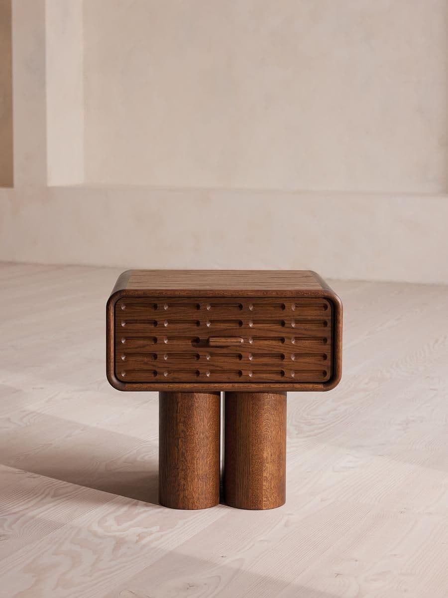 A modern wooden nightstand featuring a rectangular body with rounded corners and a slatted front panel, suggesting drawer or door access. It stands on two thick cylindrical legs. The wood exhibits a warm, medium-brown grain. — 3D model