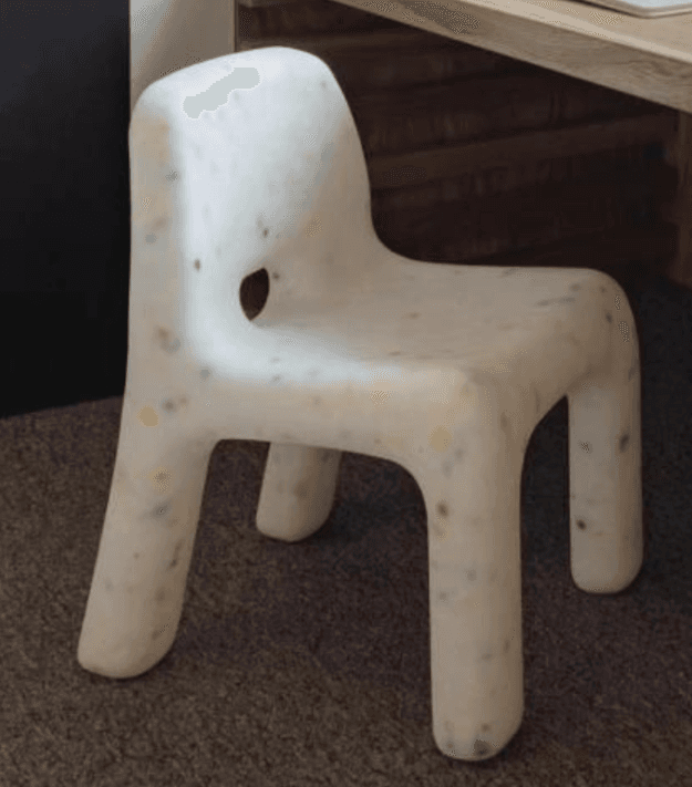 A small, monolithic children's chair made from recycled white plastic with multi-colored flecks. It features a low backrest with a cutout and thick, integrated legs, designed for stability and durability. — 3D model