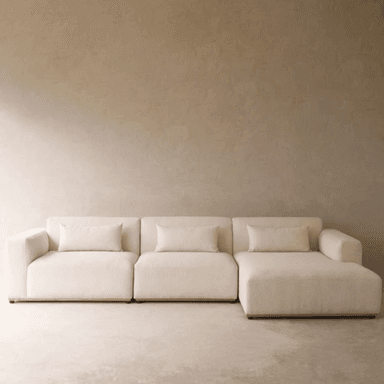 A cream-colored, modern modular sectional sofa composed of multiple units, including a chaise lounge. The sofa features a low profile, straight lines, block arms, and is upholstered in a textured fabric. It is accompanied by matching rectangular throw pillows. — 3D model