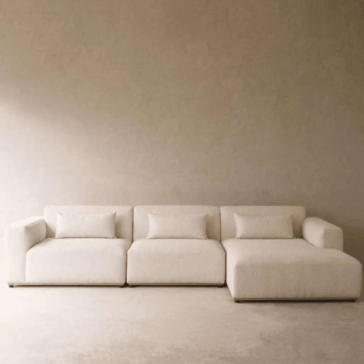 A cream-colored, modern modular sectional sofa composed of multiple units, including a chaise lounge. The sofa features a low profile, straight lines, block arms, and is upholstered in a textured fabric. It is accompanied by matching rectangular throw pillows. — 3D model