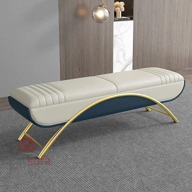An elongated, contemporary bench featuring a two-tone upholstered top and a distinctive curved gold-finished metal base. The main seating surface is a light cream or beige, detailed with parallel channel tufting, while the side panels of the cushion are upholstered in a contrasting dark blue. The metal base forms a graceful arc beneath the bench, with additional support bars. The design suggests a modern and elegant aesthetic. — 3D model