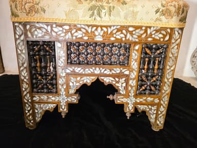 A highly ornate accent chair featuring intricate marquetry and inlay work. The chair has a tall, elaborately carved back with floral motifs and geometric patterns. The seat is upholstered in a light-colored fabric with a floral pattern. The base of the chair, including the legs and the area beneath the seat, is constructed from dark wood and heavily decorated with mother-of-pearl inlay, creating floral and geometric designs. The legs have an arched structure. The overall construction suggests a piece of antique or artisan craftsmanship. — 3D model