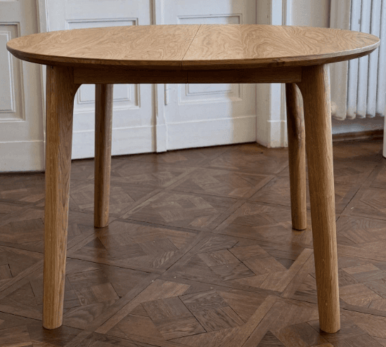 A round-topped side table with three tapered legs. The tabletop has a visible wood grain pattern and is light brown in color. The legs are also made of wood and are angled outwards from the center. — 3D model