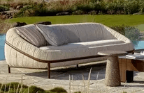 A modern outdoor sofa with a distinctive curved silhouette, upholstered in light-colored, channel-tufted fabric, featuring a dark contrasting linear frame and throw pillows. — 3D model