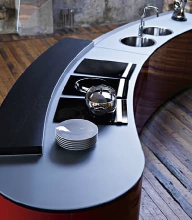 A modern, curved kitchen island featuring a rich wood veneer base and a light grey solid surface or laminate countertop. The island is equipped with three integrated black ceramic cooktops, two stainless steel circular sinks, and a single gooseneck faucet. The design is sleek and contemporary, with a minimalist aesthetic and functional integration of kitchen appliances. — 3D model