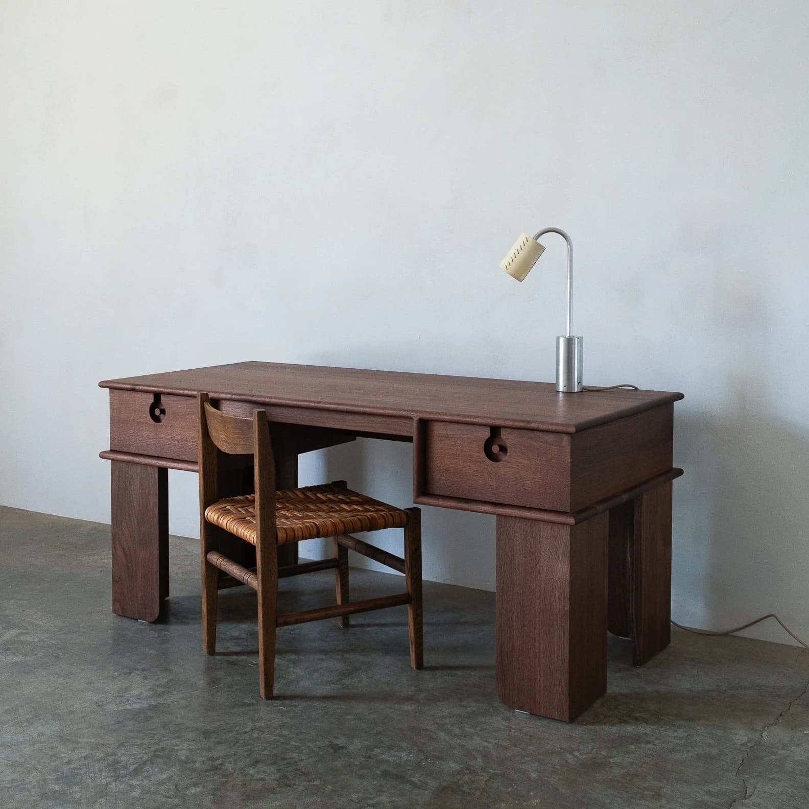 A dark wood desk with a simple, geometric design. It features a rectangular top with two drawers on either side, each with a circular, carved handle integrated into the drawer face. The desk is supported by four thick, rectangular legs. A wooden chair with a woven seat is positioned in front of the desk, and a small, modern desk lamp with a chrome finish is placed on the right side of the desk's surface. — 3D model