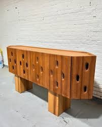 A long, low credenza with a unique, carved front featuring irregularly shaped cutouts. The piece rests on two thick, rectangular wooden legs. The top surface is smooth and appears to be a single, solid piece of wood. The overall design is modern with a natural, organic feel. — 3D model