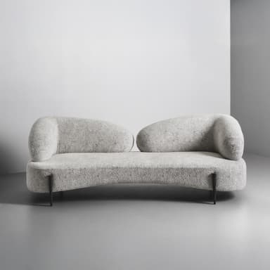A close-up view of a segment of a modern sofa, featuring rounded, organic lines and textured upholstery. The sofa has a plush appearance, with a low profile and substantial cushioning. A single, distinctly designed metal leg is visible, supporting the rounded front base of the sofa. The leg has a dark, textured finish with a cap that appears to be a different material, possibly metal or wood with a metallic finish. — 3D model