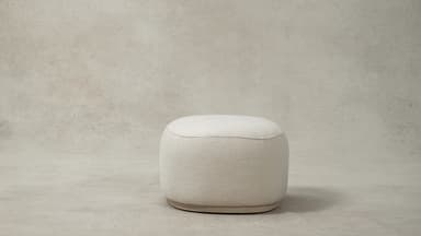 A contemporary upholstered ottoman with a rounded oval shape, covered in a plush cream bouclé fabric. The design features soft, curvilinear forms and a low-profile base, contributing to its modern and inviting aesthetic. A subtle seam runs along the perimeter of the top surface, connecting to vertical seams on the sides. — 3D model