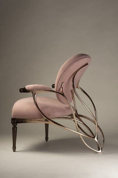 A contemporary lounge armchair featuring a dramatic sculptural frame crafted from gilded metal, supporting a plush, blush pink velvet upholstered seat and backrest. The chair's design combines soft, inviting textures with bold, curvilinear metallic elements, creating a visually striking piece. It rests on four dark, turned wooden legs with subtle metallic caps. — 3D model