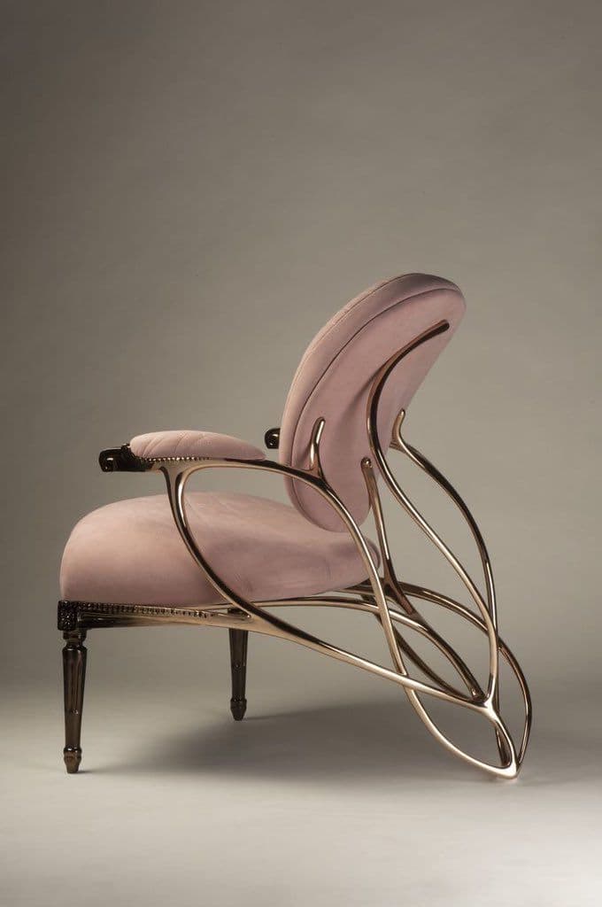 A contemporary lounge armchair featuring a dramatic sculptural frame crafted from gilded metal, supporting a plush, blush pink velvet upholstered seat and backrest. The chair's design combines soft, inviting textures with bold, curvilinear metallic elements, creating a visually striking piece. It rests on four dark, turned wooden legs with subtle metallic caps. — 3D model