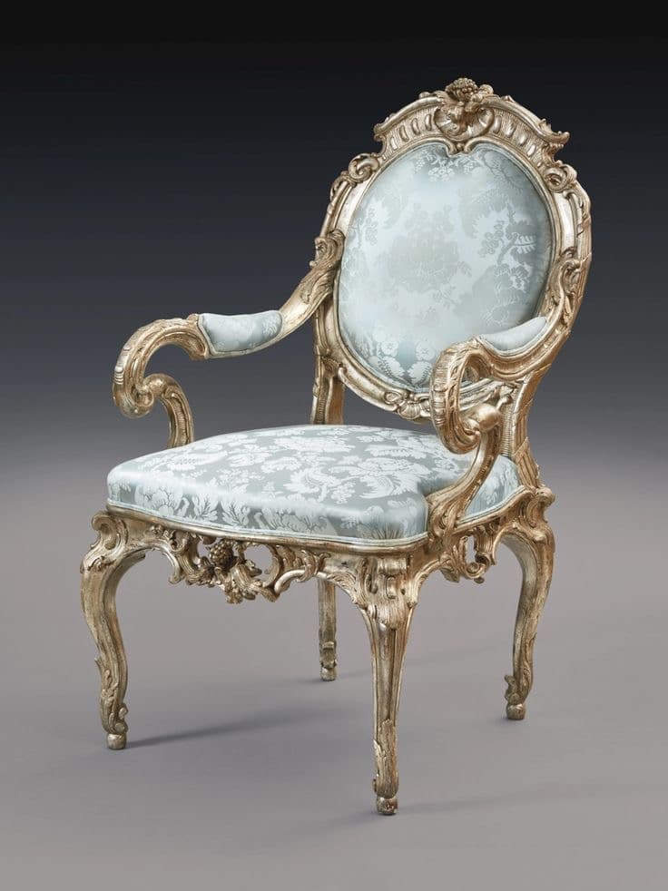 An ornate armchair with a carved wooden frame and upholstered seat and back. The frame features elaborate scrollwork and floral motifs, finished in a distressed gold or silver leaf effect. The upholstery is a light blue fabric with a damask pattern. The chair has cabriole legs and curved arms. — 3D model