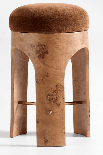 A contemporary stool featuring a round, plush brown upholstered seat set upon a distinctive burl wood veneer base. The base forms a continuous, curved structure with arched cutouts, creating a modern and sculptural silhouette. A brass bar serves as a footrest, extending between two of the wooden supports. — 3D model