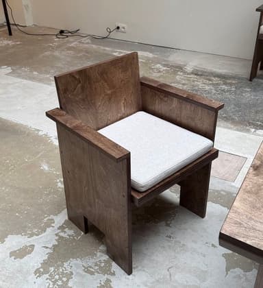 A modern, angular armchair constructed from dark-stained wood panels, featuring a boxy silhouette, wide flat armrests, and a separate light gray upholstered seat cushion. — 3D model