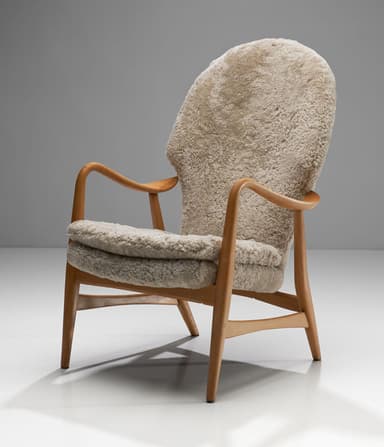 A high-backed armchair with a wooden frame and plush sheepskin upholstery. The frame consists of curved wooden arms and legs. The seat and back are fully upholstered in a textured, cream-colored sheepskin material, giving it a soft and inviting appearance. The design suggests a focus on comfort and organic forms. — 3D model