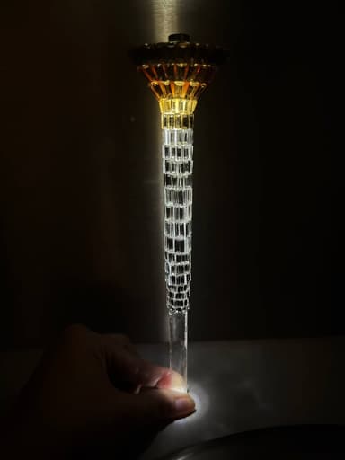 A tall, slender decorative object, likely functioning as a lamp, featuring a prominent faceted clear crystal stem. The top part consists of an amber-colored, fluted or pleated glass shade, capped with a dark, dome-shaped metallic element. A golden-toned band separates the shade from the main body. The stem is composed of numerous clear, rectangular crystal segments arranged in a stacked, tapering formation, culminating in a smooth, clear cylindrical base. The overall design suggests a luxurious and elaborate aesthetic. — 3D model