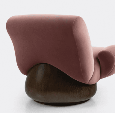 A plush armchair with a rounded silhouette and voluminous cushioning. The seat and backrest are upholstered in a soft, rose-colored velvet fabric. It features integrated armrests that flow seamlessly from the main body of the chair. The chair is supported by a substantial, dark-stained wooden base with a rounded, drum-like shape. — 3D model