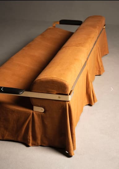 A long, rectangular sofa with a curved bolster backrest. The sofa is upholstered in a textured, light brown fabric. It features a metal frame with armrests that have black accents. The base of the sofa is covered by a draped skirt of the same fabric, creating a segmented appearance. — 3D model