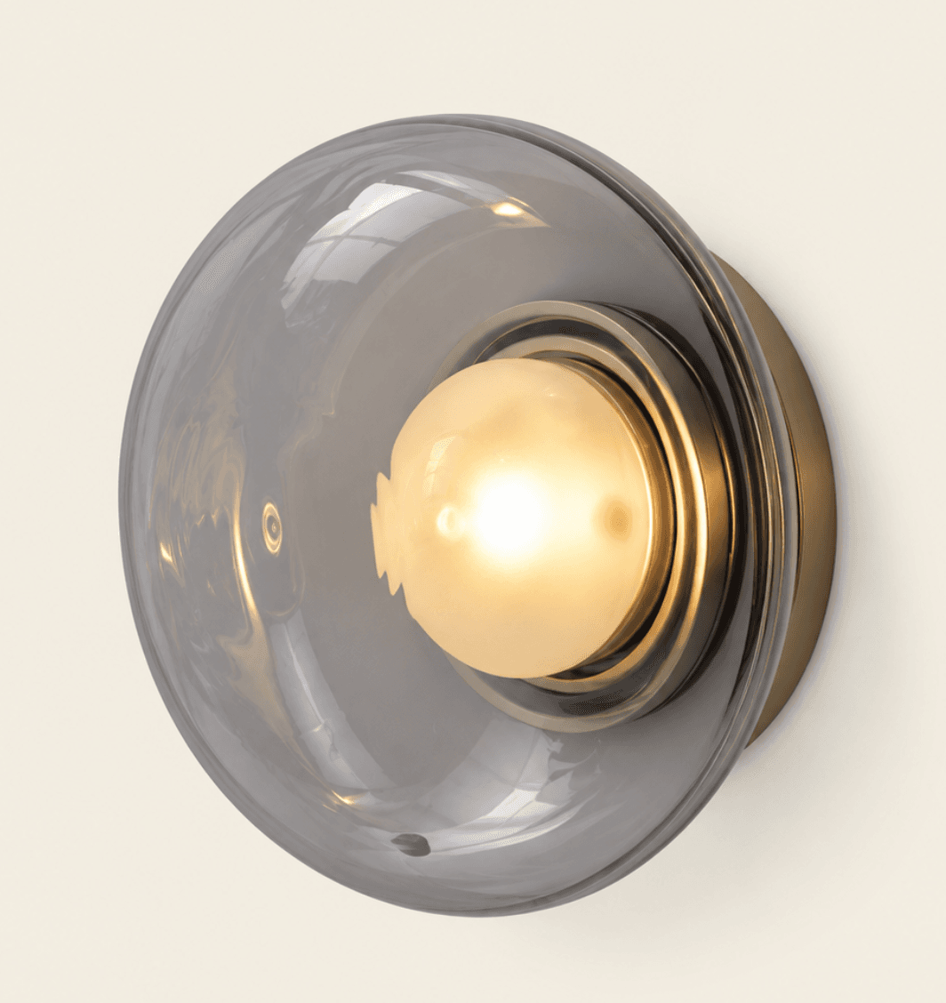 A modern wall sconce featuring a semi-spherical smoky gray glass shade encasing a warm-toned bulb, mounted on a brushed brass circular backplate. — 3D model