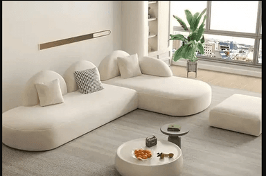 A cream-colored, low-profile modular sectional sofa featuring an integrated chaise lounge. The sofa is characterized by its soft, organic rounded shapes and is upholstered in a textured boucle fabric. It includes several small lumbar pillows, one of which has a black and white checkered pattern. — 3D model