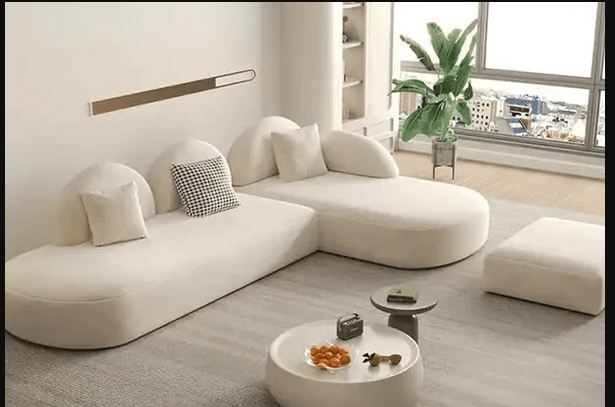 A cream-colored, low-profile modular sectional sofa featuring an integrated chaise lounge. The sofa is characterized by its soft, organic rounded shapes and is upholstered in a textured boucle fabric. It includes several small lumbar pillows, one of which has a black and white checkered pattern. — 3D model
