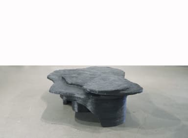 A sculptural, low-lying table featuring an organic, multi-tiered design. The dark, matte material has a layered texture, mimicking natural rock formations or topographical maps. The table top comprises several irregularly shaped, overlapping planes at varying heights, supported by an equally irregular and textured base. — 3D model