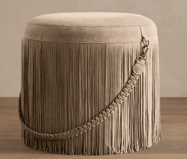 A round, upholstered ottoman featuring a full skirt of long fringe. The top surface is smooth and upholstered in a suede-like material. A decorative braided strap, also in the same material, is draped around the side, secured by a metal clasp with an antique brass finish. The fringe extends to the floor, creating a cohesive and textured look. — 3D model