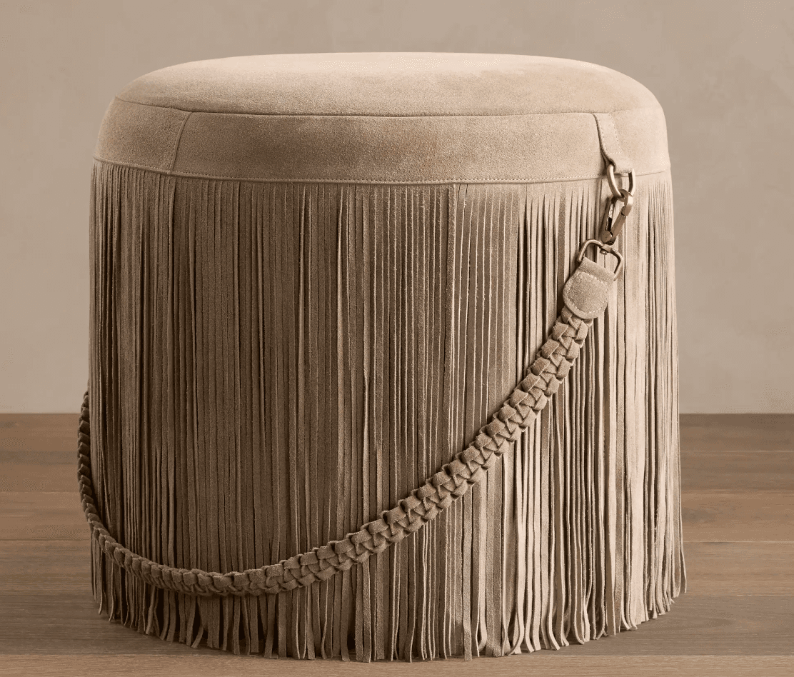 A round, upholstered ottoman featuring a full skirt of long fringe. The top surface is smooth and upholstered in a suede-like material. A decorative braided strap, also in the same material, is draped around the side, secured by a metal clasp with an antique brass finish. The fringe extends to the floor, creating a cohesive and textured look. — 3D model