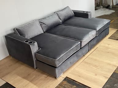 A modern sectional sofa with a chaise lounge extension on one side. The sofa features multiple seat cushions and back cushions, upholstered in a soft, plush gray fabric. It has a piped trim in a lighter contrasting color along the edges of the cushions and frame. Two integrated cup holders are visible on the armrests. The base appears to be a solid structure, possibly with decorative metal feet. — 3D model
