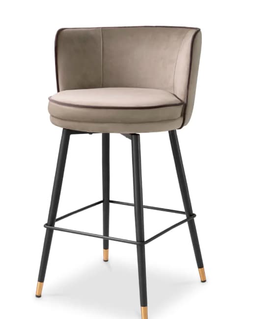 An elegant contemporary bar stool featuring a comfortable upholstered bucket seat in a light beige or taupe fabric, likely velvet, with contrasting darker piping. The seat swivels and is supported by a sturdy black metal frame with four tapered legs. Each leg is accented with a polished brass cap at the foot, and a circular metal footrest is integrated into the base. — 3D model