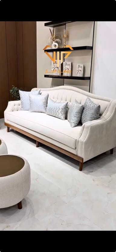 A tufted sofa with a curved back and a wooden frame. The sofa is upholstered in a light-colored fabric and comes with four decorative throw pillows in various silver and grey textured patterns. The base has visible wooden legs and a complementary wooden trim along the bottom of the upholstered frame. — 3D model