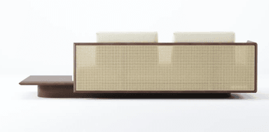 A low-profile sofa featuring a wooden base with an extended side surface. The seating area is upholstered in a textured cream fabric. It is equipped with two large back cushions and two smaller cylindrical bolster cushions. The back of the sofa incorporates woven cane panels framed by dark wood. — 3D model