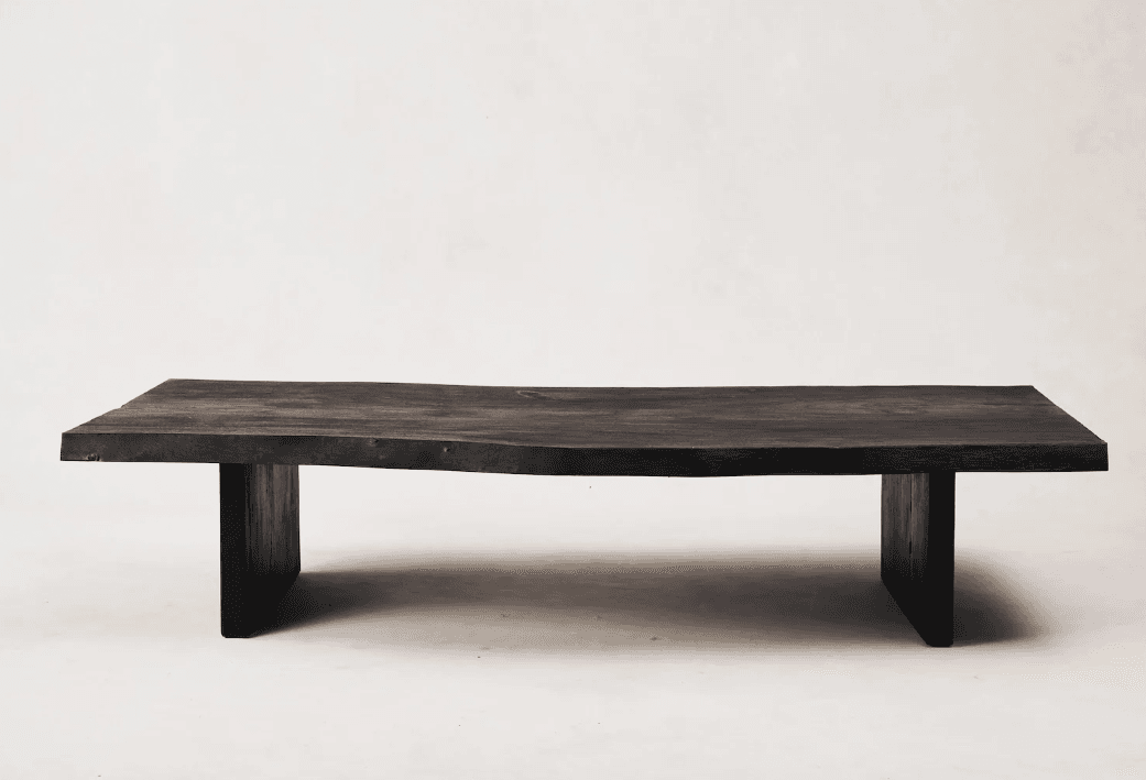 A low, rectangular coffee table with a live-edge top and thick, blocky legs. The tabletop has an irregular, natural edge on one side, and the surface displays visible wood grain. The legs are positioned inset from the ends of the tabletop. — 3D model