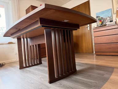 A modern dining table featuring a dark brown wooden top with elegantly curved edges and a matching slatted base. The table top displays a visible wood grain pattern, suggesting a veneer finish, and shows some minor surface wear. The base appears robust, designed with vertical wooden slats, which likely form a pedestal or trestle-style support. — 3D model